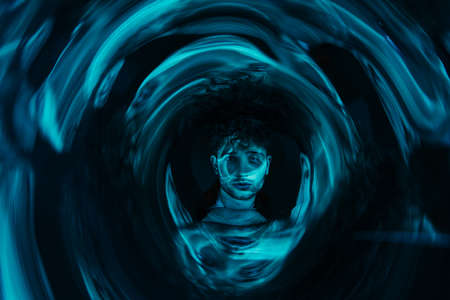 curly man looking at camera through futuristic neon blue circle on dark backgroundの写真素材