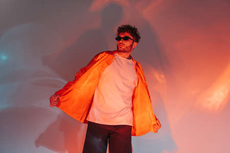 curly and trendy man in sunglasses and orange shirt posing in studio with lightingの写真素材