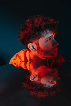 reflection in mirror of curly and stylish man in orange shirt posing on blue