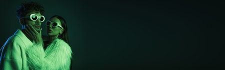 fashionable woman in sunglasses and trendy outfit touching face of man on dark blue, bannerの写真素材