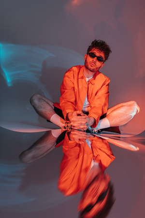 full length of stylish man in sunglasses posing while sitting in studio with colorful light