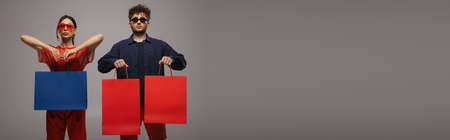 young couple in trendy outfits and sunglasses holding shopping bags isolated on grey, bannerの写真素材