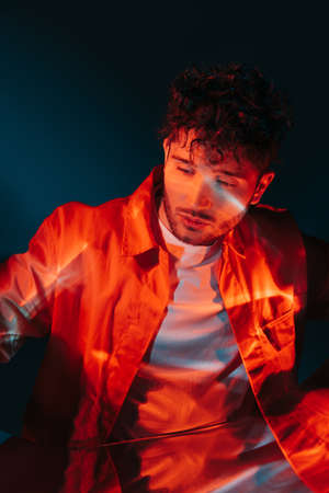 lighting on face of curly and stylish man in orange shirt posing on blueの写真素材