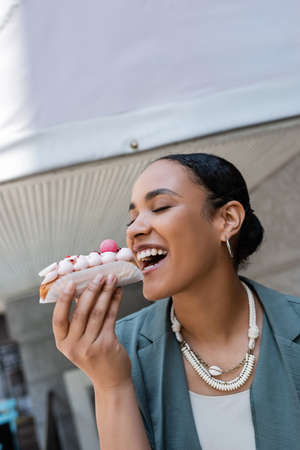 Young african american woman biting eclair near confectionery outdoorsの写真素材