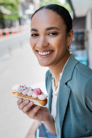 Pretty young customer holding delicious eclair and looking at camera on terrace of cafeの写真素材