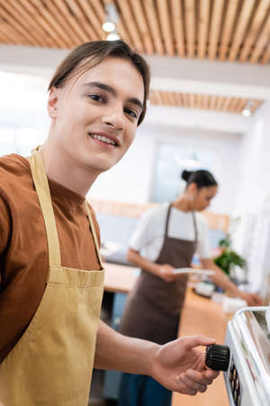 Smiling barista in apron looking at camera near coffee machine in sweet shopの写真素材