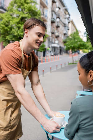 Positive salesman putting cappuccino near african american client on terrace of sweet shopの写真素材