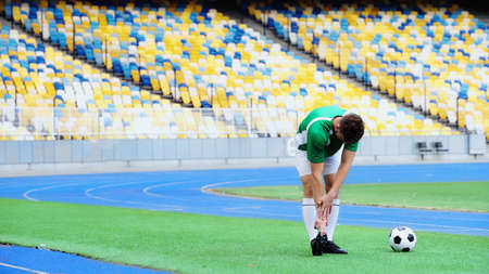 young football player in uniform stretching near ball of green field