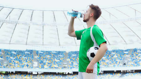 side view of young football player in green t-shirt holding ball while drinking waterの写真素材