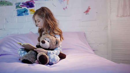 Side view of kid in pajama reading book and holding soft toy on bed at homeの写真素材