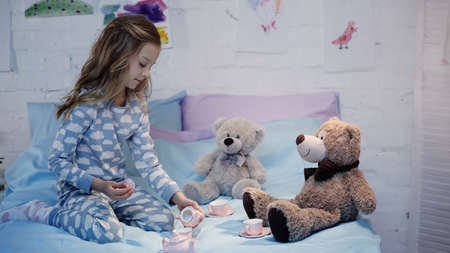 Preteen kid in pajama playing with tea cups near toys on bedの写真素材