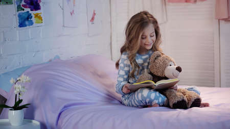 Smiling preteen child in pajama reading book near soft toy on bed in eveningの写真素材