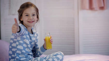 Smiling kid in pajama showing thumb up and holding orange juice on bedの写真素材