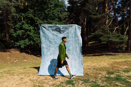 side view of african american man in green blazer and black shorts walking near blue cloth in park
