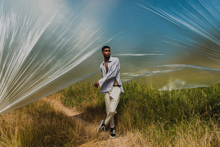 full length of african american man in summer outfit standing under polyethylene film in grassy...