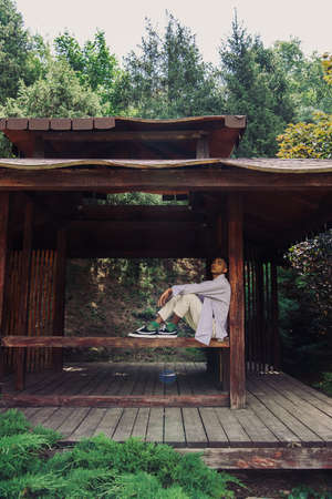 full length of african american man in summer clothes and sneakers sitting in wooden gazeboの写真素材