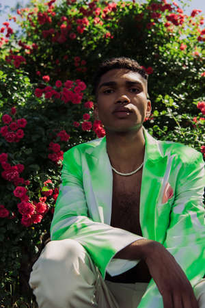 african american man in beads and trendy blazer looking at camera near blossoming bushes