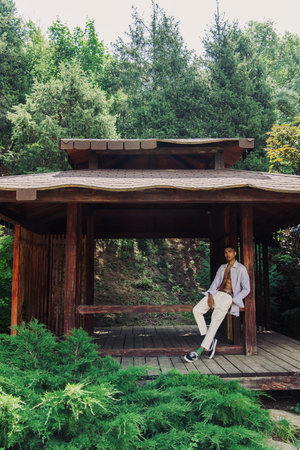 full length of trendy african american man in wooden gazebo in green parkの写真素材