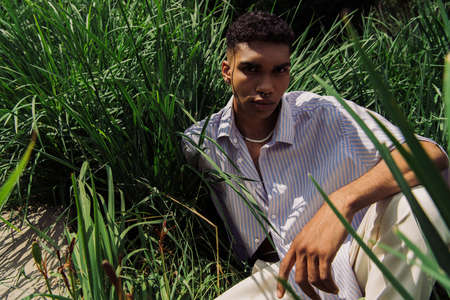high angle view of young african american man in trendy shirt sitting on grass and looking at cameraの写真素材