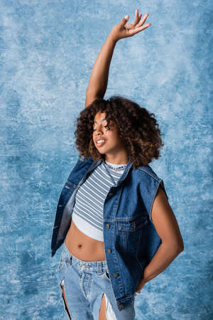 african american woman in denim vest and crop top standing with closed eyes and raised hand on blue...