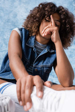 brunette african american woman in denim vest obscuring face with hand on blue backgroundの写真素材