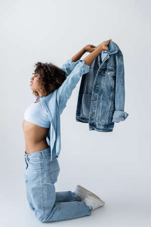 side view of african american woman holding denim jacket while standing on knees on grey background