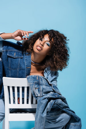 trendy african american woman in denim clothing looking at camera on chair isolated on blueの写真素材
