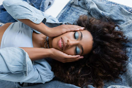 top view of african american woman with makeup touching face while lying on denim garments on grey...