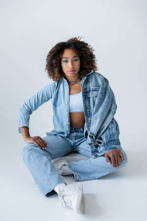 curly african american woman in blue jeans and sneakers sitting on grey backgroundの写真素材