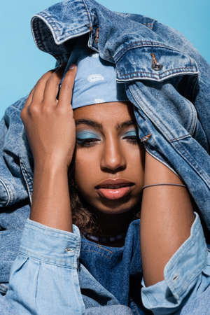 african american woman with makeup covering head with denim jacket isolated on blueの写真素材
