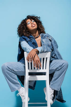 trendy african american woman in denim clothes posing on chair with closed eyes on blue backgroundの写真素材