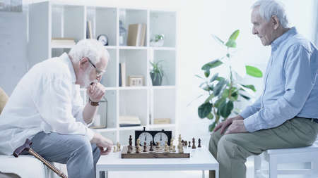 side view of thoughtful man playing chess with senior friend in living roomの写真素材