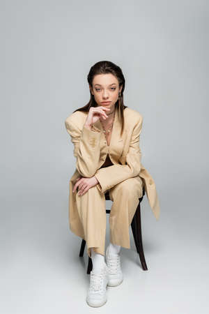 full length of stylish woman in trendy outfit looking at camera while sitting on chair on greyの写真素材