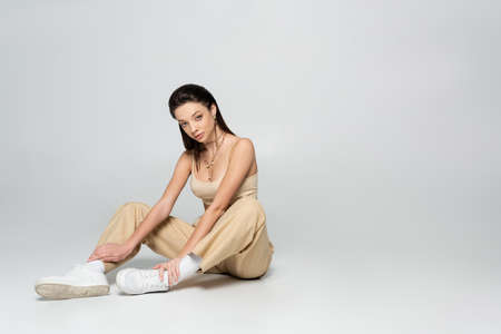 full length of brunette woman in stylish beige outfit and trendy accessories sitting on greyの写真素材