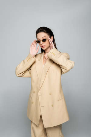 young woman in stylish beige outfit wearing trendy sunglasses isolated on greyの写真素材