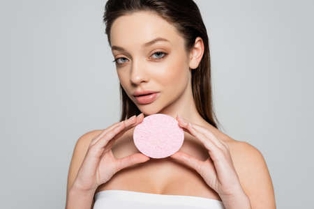 brunette young woman with bare shoulders holding exfoliating sponge isolated on greyの写真素材