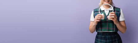 Cropped view of cheerful student in uniform holding wireless headphones isolated on purple, bannerの写真素材