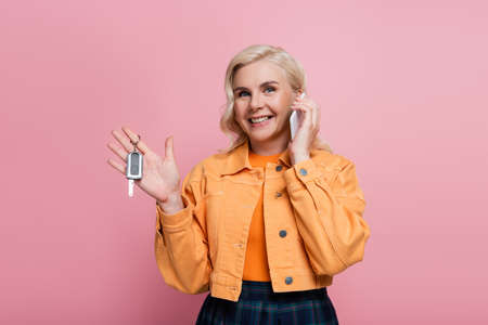 Positive woman in jacket talking on smartphone and holding car key isolated on pink