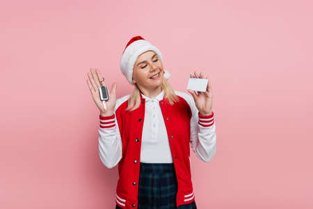 Cheerful woman in santa hat holding car key and empty driving license isolated on pink