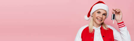 Positive woman in santa hat and baseball jacket holding car key isolated on pink, banner