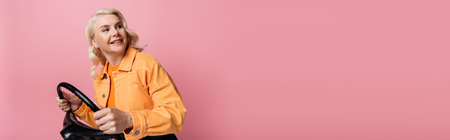 Smiling blonde driver in jacket holding steering wheel ad looking away isolated on pink, bannerの写真素材