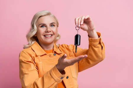 Positive blonde woman holding car key isolated on pink