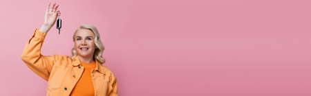 Cheerful blonde driver in jacket holding car key isolated on pink, banner