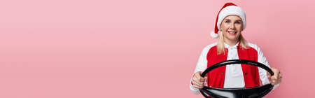 Positive woman in santa hat holding steering wheel isolated on pink, bannerの写真素材