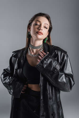 sensual woman in leather clothes and silver necklaces posing with closed eyes isolated on greyの写真素材