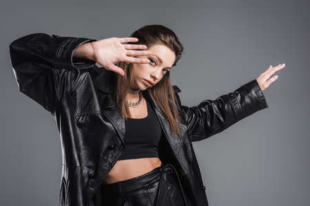 young and stylish woman in black leather coat gesturing and looking at camera isolated on greyの写真素材