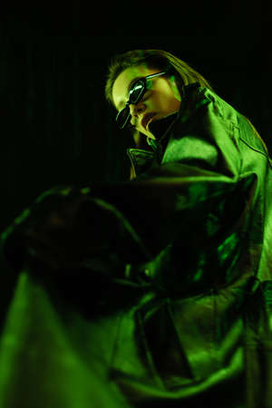 low angle view of woman in leather coat and dark sunglasses in green light isolated on blackの写真素材