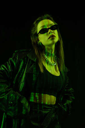 trendy woman in sunglasses and silver necklaces posing with hands in pockets in green light isolated on blackの写真素材