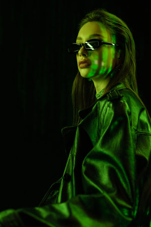 young brunette woman in leather coat and sunglasses in green neon light isolated on black