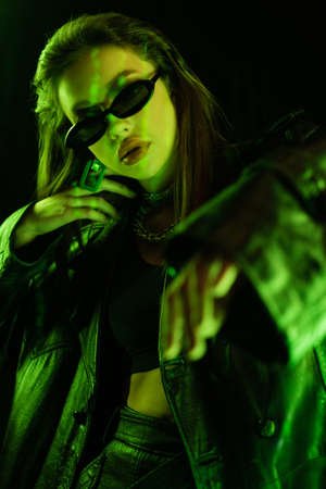 trendy woman in dark sunglasses and leather coat gesturing in green light isolated on blackの写真素材